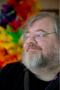 Tom Smith: A man with salt and pepper hair and beard, wearing glasses and a black tshirt looks toward the left.  Behind him are rainbow-colored flowers.