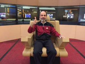 A smiling man in a zip-up jacket in the style of a Starfleet uniform (Next Generation era) sits in a replica of Captain Kirk's chair on a bridge set